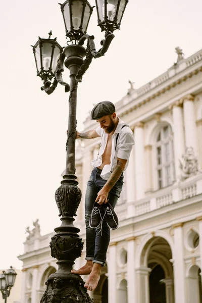 Young bearded tattooed man. A romantic guy in a white shirt, cap and suspenders walks in the city. revolves around a lamppost. Peaky Blinders. old-fashioned, retro.