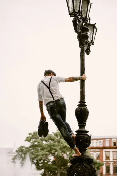 Young bearded tattooed man. A romantic guy in a white shirt, cap and suspenders walks in the city. revolves around a lamppost. Peaky Blinders. old-fashioned, retro.