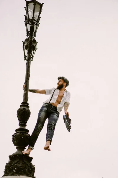 Young bearded tattooed man. A romantic guy in a white shirt, cap and suspenders walks in the city. revolves around a lamppost. Peaky Blinders. old-fashioned, retro.
