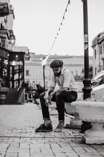 Young bearded tattooed man. A romantic guy in a white shirt, cap and suspenders walks in the city. revolves around a lamppost. Peaky Blinders. old-fashioned, retro.