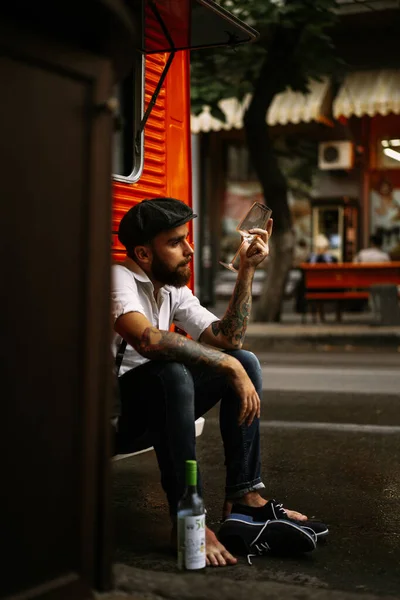 Young bearded tattooed man. A romantic guy in a white shirt, cap and suspenders walks in the city. revolves around a lamppost. Peaky Blinders. old-fashioned, retro.