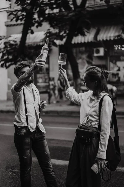 A young man in a white shirt and a cap with a woman on the street drinks wine from glasses. Friends meeting