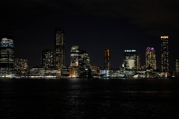 New York City Manhattan downtown at night with skyscrapers illuminated over the Hudson River with reflections.