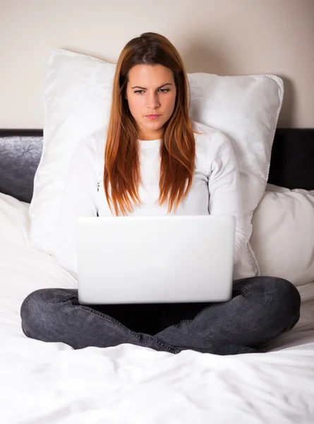 Young woman using her laptop while sitting on her bed — Stock Photo, Image