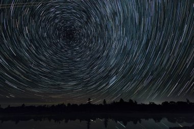 Yıldızlı gece yıldız izleri bir göl ve orman üzerinde, Kutup yıldızları etrafında hareket ediyor.