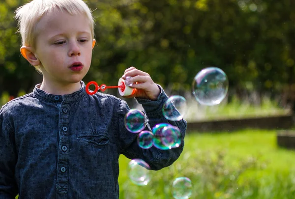 En liten pojke blåser såpbubblor i trädgården på en sommardag — Stockfoto