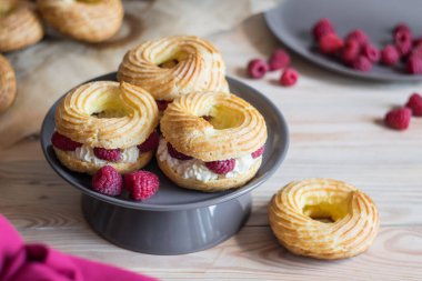 Kremalı poğaça halkaları, eklair choux, gri tabakta taze ahududu ile süslenmiş, seçici bir odak noktası.