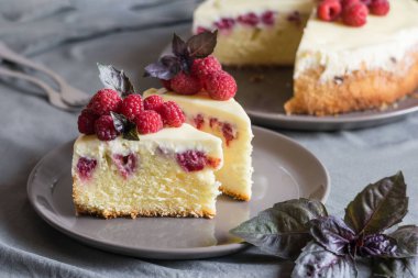 Ahudududulu cheesecake dilimleri, taze ahududu ve bazil yapraklarıyla gri tabakta, arka planda bütün peynirli kek. Seçici odak