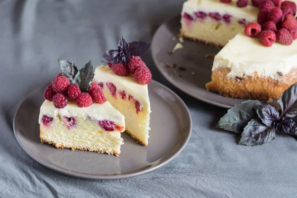 Ahudududulu cheesecake dilimleri, taze ahududu ve bazil yapraklarıyla gri tabakta, arka planda bütün peynirli kek. Seçici odak