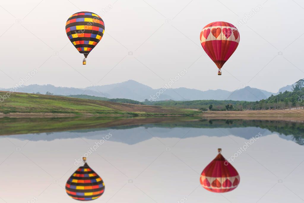 Photo of colorful aerostat balloons with basket floating in the air above lake with beautiful scenery behind them