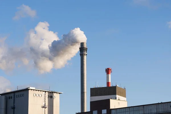 Industrial chimney with smoke and factory buildings against the blue sky — Stock Photo, Image