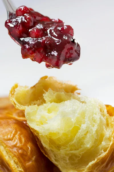 Red cranberry jam on a spoon above a fresh croissant, breackfast closeup, vertical — Stock Photo, Image