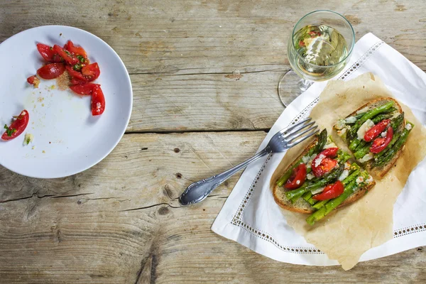 Green asparagus, tomatoes and parmesan on toasted bread and tomato salad, rustic wood, copy space, from above — Stock Photo, Image