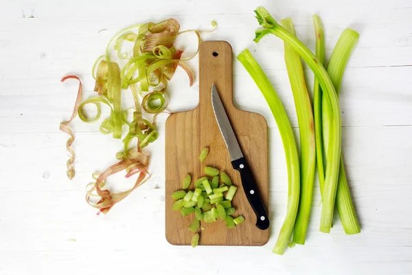 Tallos de ruibarbo y piezas en rodajas, cuchillo de cocina y tabla de cortar en madera blanca, desde arriba —  Fotos de Stock