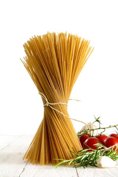 Stelletje volkoren spaghetti staande op wit hout met sommige tomaten en kruiden tegen een witte achtergrond — Stockfoto