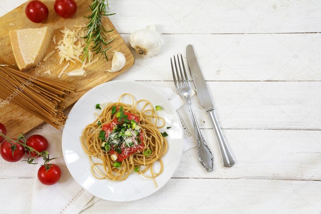 whole wheat spaghetti with fresh tomato sauce and a kitchen board with some ingredients on a white wooden background 