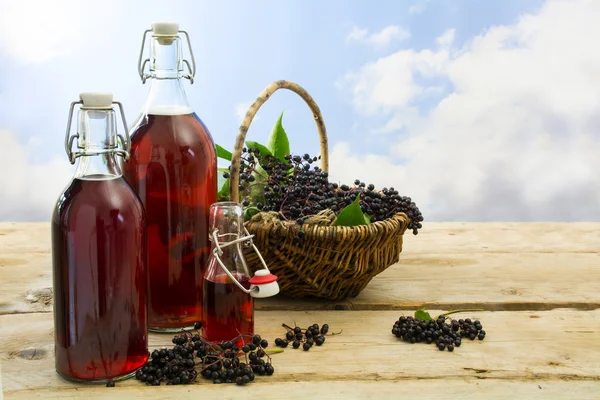 Black elderberry juice in bottles and a basket with fresh fruits on rustic wood against a blue sky — Stock Photo, Image