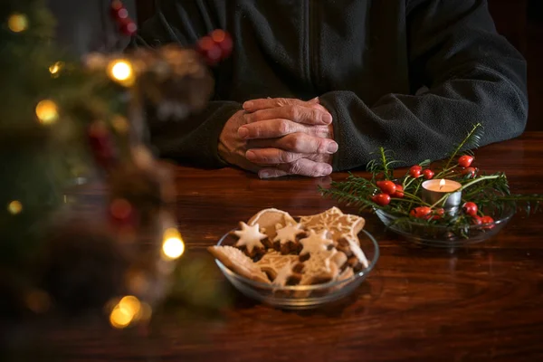 Praying hands of an elderly man on a table with Christmas cookies, burning candle and festive decoration, lonely holidays during the coronavirus pandemic, copy space, selected focus, narrow depth of field