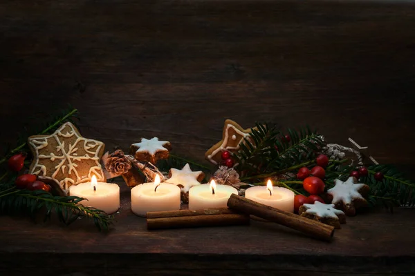 Four small candles lit for Advent and Christmas decoration like gingerbread cookies, branches and rose hips on a dark rustic wooden background, generous copy space, selected focus