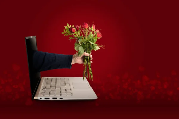 Hand of a man with a bouquet of roses coming out of a laptop screen, digital love on Valentines day with social distance during covid-19 pandemic, red background with heart shape bokeh, copy space