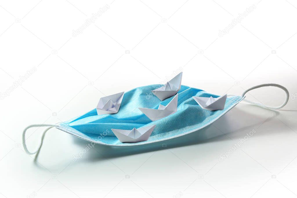 Paper boats with different courses sail on a surgical face mask like on the high seas, metaphor for searching a way out of the coronavirus crisis or dangerous tourism during covid-19 pandemic, copy space, selected focus