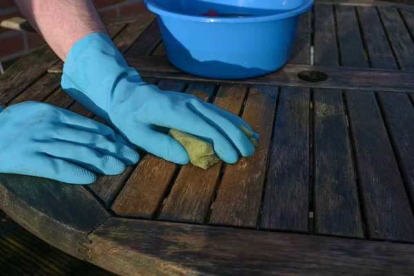Hands in blue rubber gloves cleaning a weathered wooden garden table at the beginning of spring forthe outdoor season, copy space, selected focus