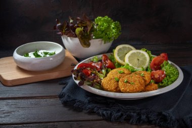 Nohuttan kızartılmış falafel, susam tohumları, baharatlar ve otlar, salata ve yoğurt sosu ile servis edilmiş, kırsal bir ahşap masa üzerinde koyu bir arka plan, kopyalama alanı, seçilmiş alan, dar bir alan derinliği