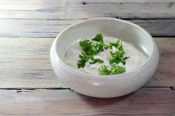 Yogurt dip with herbs and parsley garnish in a small ceramic bowl on a light rustic wooden table, copy space, selected focus, narrow depth of field