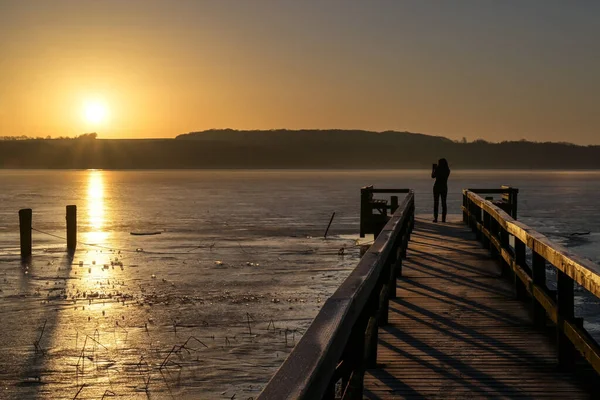 凍った湖の上に昇る太陽の写真を撮る木製の桟橋のシルエットとして後ろから若い女性 コピースペース 選択された焦点 — ストック写真