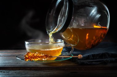 Sıcak buharlı çay demlikten cam fincana dökülür, bir çubuk kaya şekeri, hareket bulanıklığı, fotokopi alanı ile koyu kırsal ahşap arka plan, seçilmiş odak, dar alan derinliği ile servis edilir.