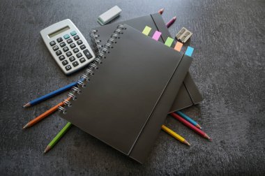 Siyah spiral defterler, renkli boyalar, hesap makinesi ve sabit malzemeler koyu gri bir masa üzerinde, kopyalama alanı, seçili odak, dar alan derinliği