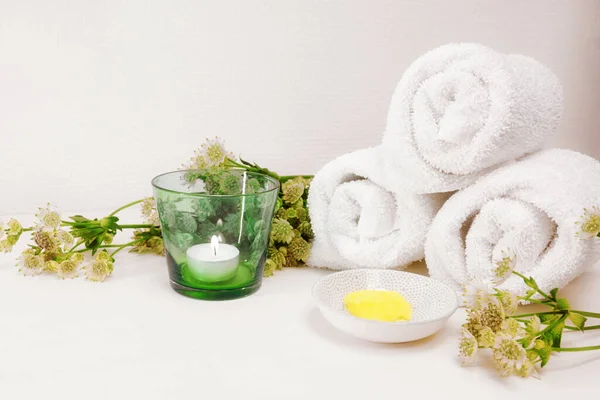 Cosmetic moisturizer or emollient for pedicure and soft foot skin, white towels, candle and some green flowers on a light background with large copy space, selected focus, narrow depth of field