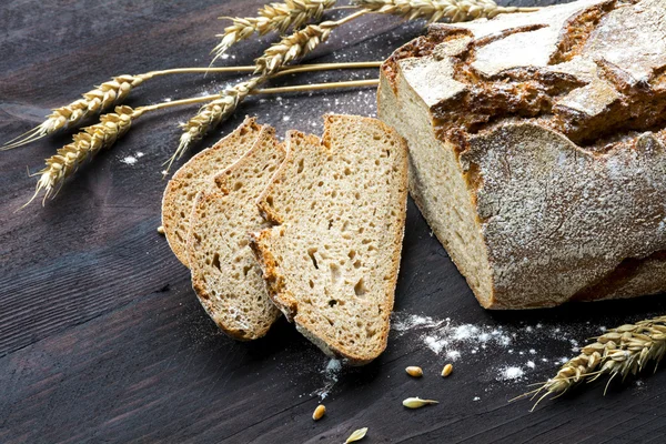 Rustic loaf of bread and slices with wheat on dark wood — Stock Photo, Image