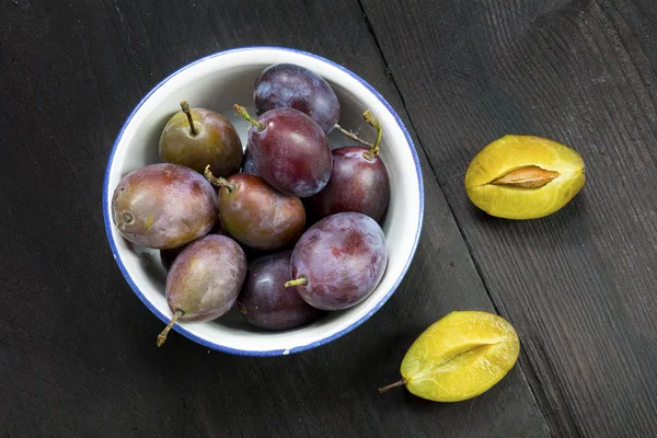 Plums in a bowl, whole and halved on dark brown wood — Stock Photo, Image