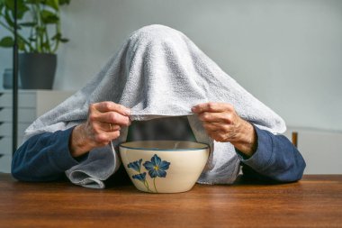 Sonbahar ve kış aylarında soğuk algınlığı ve grip mevsimi. Kafasında havluyla yaşlı bir adam sıcak bitkisel buhar soluyor. Bir kâse, sağlık konsepti, fotokopi alanı, seçilmiş odak noktası.