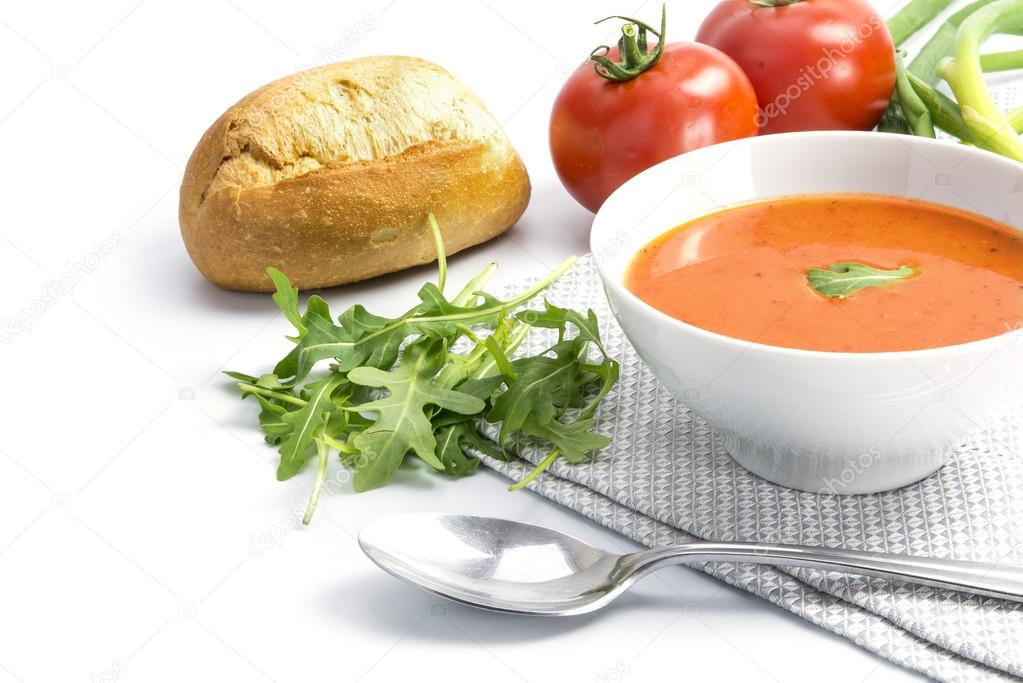 Tomato soup in a bowl with bread rolls and arugula on a white ba