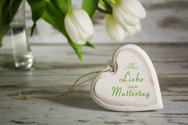 Forma de corazón de madera delante de tulipanes blancos para el día de las madres, texto alemán —  Fotos de Stock