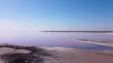 Pembe göl kıyısında mavi gökyüzü ve arka planda rüzgar türbinleri var. 