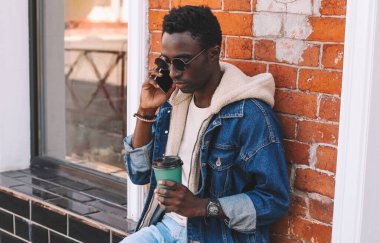 Genç bir Afrikalı 'nın portresi, elinde bir fincan kahveyle şehir caddesinde otururken akıllı telefondan konuşuyor.