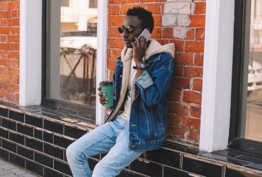 Genç bir Afrikalı 'nın portresi, elinde bir fincan kahveyle şehir caddesinde otururken akıllı telefondan konuşuyor.