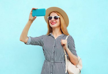 Gülümseyen genç bir kadının portresi, mavi bir arka planda hasır şapka ve sırt çantası takarak akıllı telefonuyla selfie çekiyor.