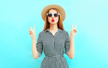 Güzel genç bir kadının portresi kırmızı dudaklarını siliyor çizgili bir elbise giyiyor, mavi arka planda yaz hasır şapkası takıyor.