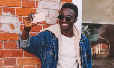 Mutlu gülümseyen Afrikalı genç bir adamın portresi. Şehir caddesinde akıllı telefonla selfie çekiyor.