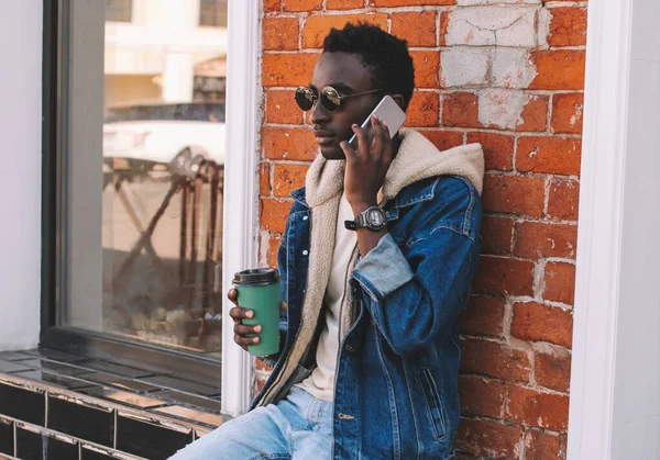 Genç bir Afrikalı 'nın portresi, elinde bir fincan kahveyle şehir caddesinde otururken akıllı telefondan konuşuyor.