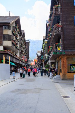 Zermatt, İsviçre - 5 Temmuz 2025: İsviçre 'nin Valais kantonunda Zermatt' ta bir dağ panoramasının önünde ahşap evler ve turistlerin bulunduğu ana alışveriş caddesi