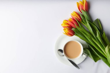 Beyaz arka planda duran kaşıklı bir fincan çay ve bir buket lale.