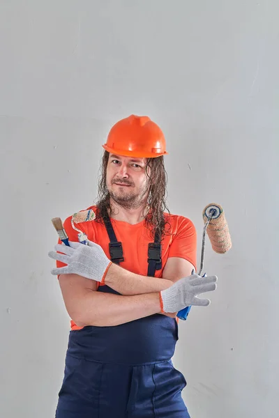 A builder stands in overalls with a roller and a brush — Stockfoto