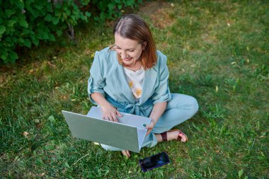 Yeşil çimenlerde oturan dizüstü bilgisayarı olan şaşırmış kadın.