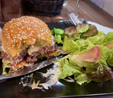 Sıcak bir restoranda siyah bir tabakta taze yeşil salata ile servis edilen, sulu etli köfte, erimiş peynir, turşu ve kremalı sos içeren gurme bir çizburgerin yakın çekimi.