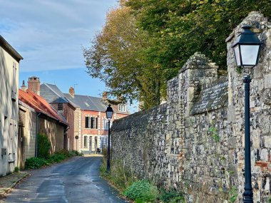 Rue des Processions, Saint-Valery-sur-Somme, taş ve kireçtaşı duvar, tuğla evler ve siyah direkler ile kaplı dar tarihi şerit, sonbahar yaprakları ve sakin mavi gökyüzü
